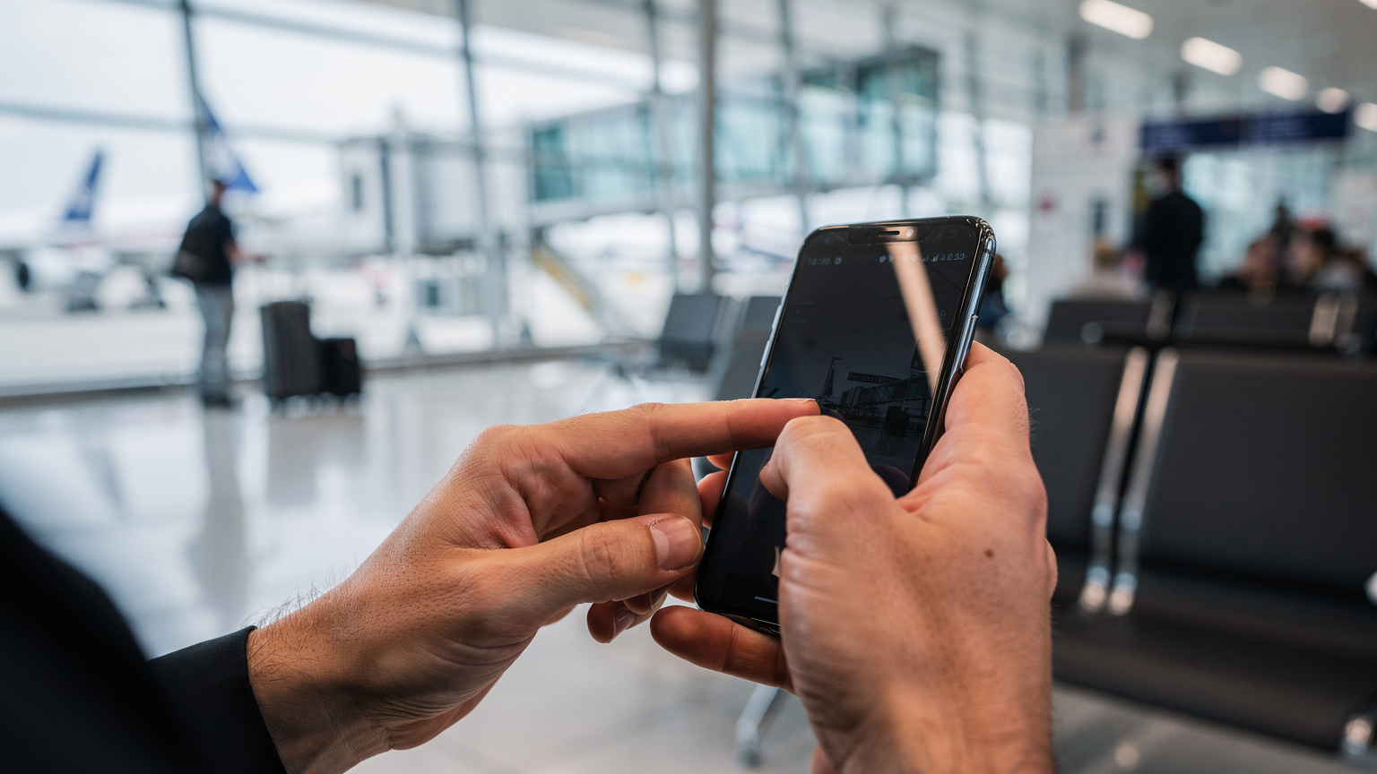 Eine Nahaufnahme zeigt die Hände eines Reisenden, der auf seinem Smartphone eine eSIM-App bedient, um sein Datenpaket zu überprüfen, während er an einem belebten Flughafen-Gate wartet.